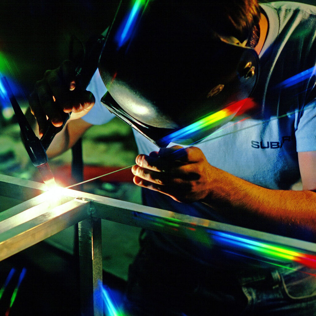 Eine Person mit einer Schweißermaske arbeitet an einem Metallgestell, während das Licht vom Schweißbrenner in bunten Farben reflektiert.