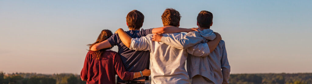 Vier junge Menschen stehen nebeneinander und halten sich gegenseitig im Arm, von hinten fotografiert.