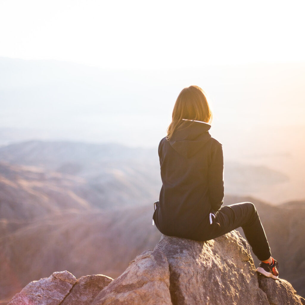 Eine junge Frau sitzt von hinten fotografiert allein auf einer Felsspitze und blickt in die Weite einer bergigen Landschaft.